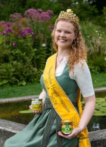 Frau mit Schärpe und Krone. Hält ein Honigglas in der Hand